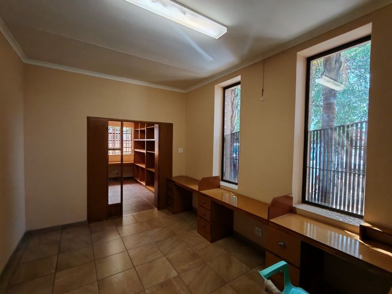 A functional office space featuring built-in wooden desks, integrated shelving, tiled flooring, and large windows with security bars.