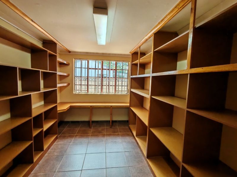 A functional utility or storage room featuring extensive built-in wooden shelving, a fixed desk area, tiled flooring, and security bars on the window.