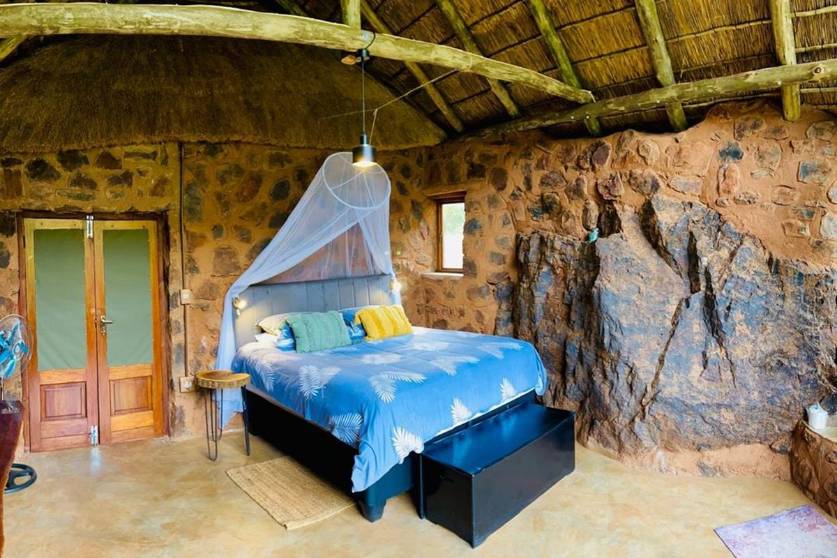 Rustic bedroom featuring exposed natural stone walls, a traditional thatched roof with timber beams, and polished concrete flooring.