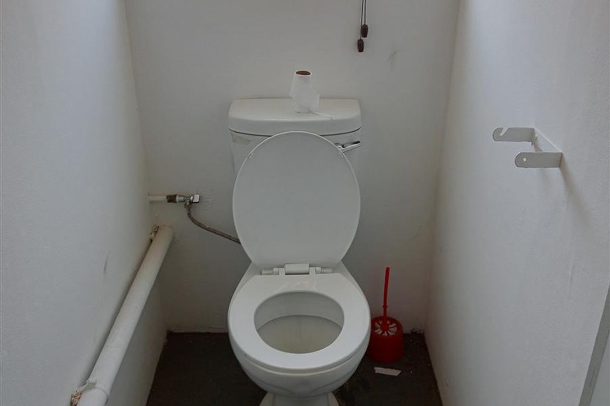A compact industrial-style restroom featuring a standard toilet, exposed plumbing, and a small window for natural light.