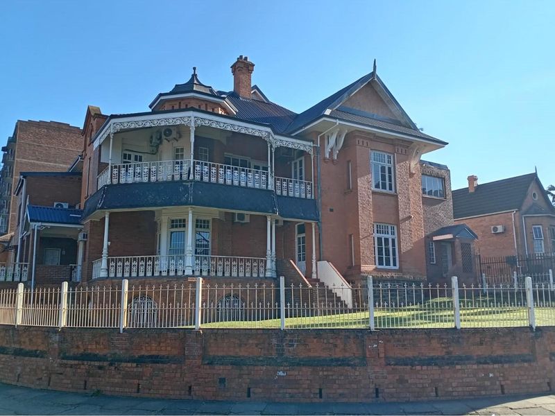 A historic multi-story commercial brick building featuring a prominent wrap-around balcony, decorative ironwork, and a secure perimeter fence.