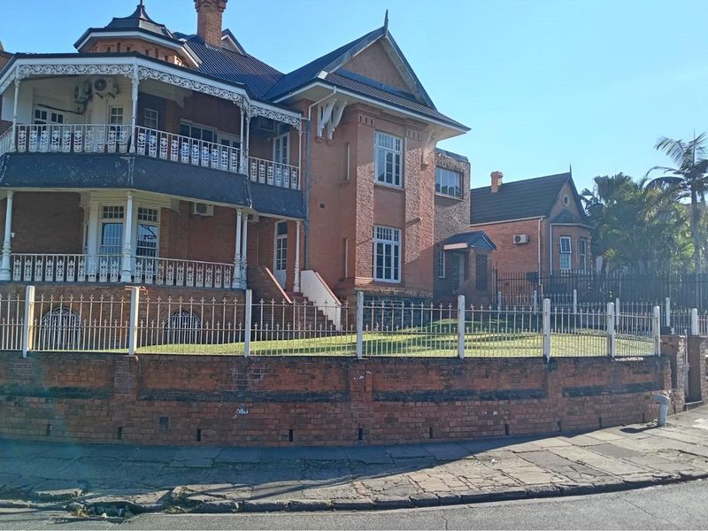 A historic multi-story brick commercial building featuring ornate metal balconies, sash windows, and a secure perimeter fence with a brick retaining wall.