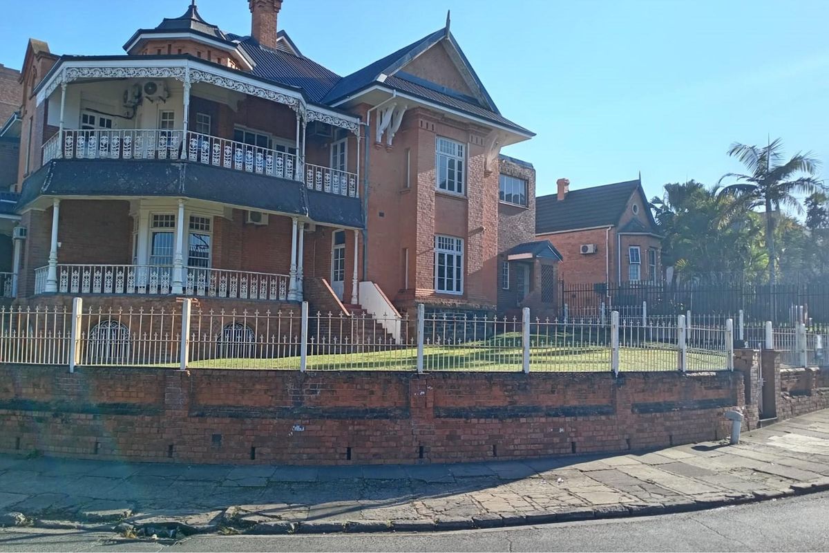 A historic multi-story brick commercial building featuring ornate metal balconies, sash windows, and a secure perimeter fence with a brick retaining wall.