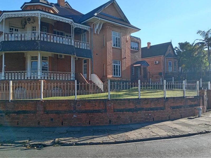 A historic multi-story commercial property featuring classic brick architecture, wrap-around balconies with decorative ironwork, and a secure perimeter fence.