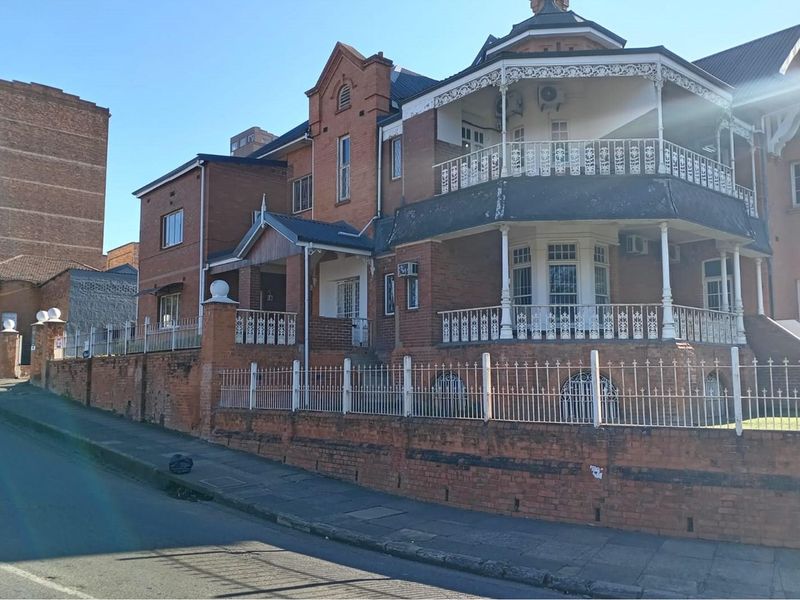 A multi-story commercial brick building featuring ornate wrought iron balconies, a pitched roof, and a secure perimeter wall along the street frontage.