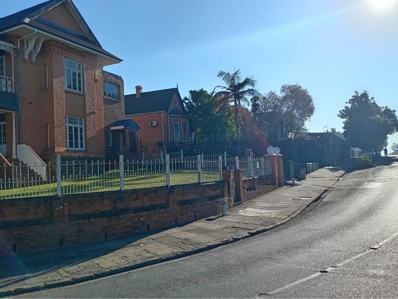 Street view of a commercial property featuring a historic brick building, perimeter fencing, and paved sidewalk access.