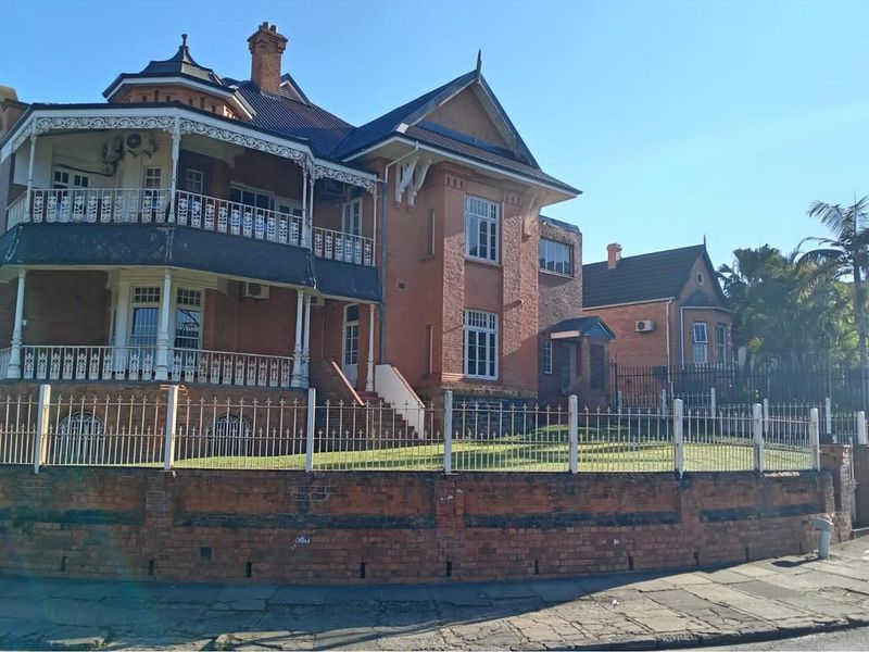 A historic commercial property featuring classic red brick architecture, a wrap-around balcony with ornate ironwork, and a secure perimeter fence.
