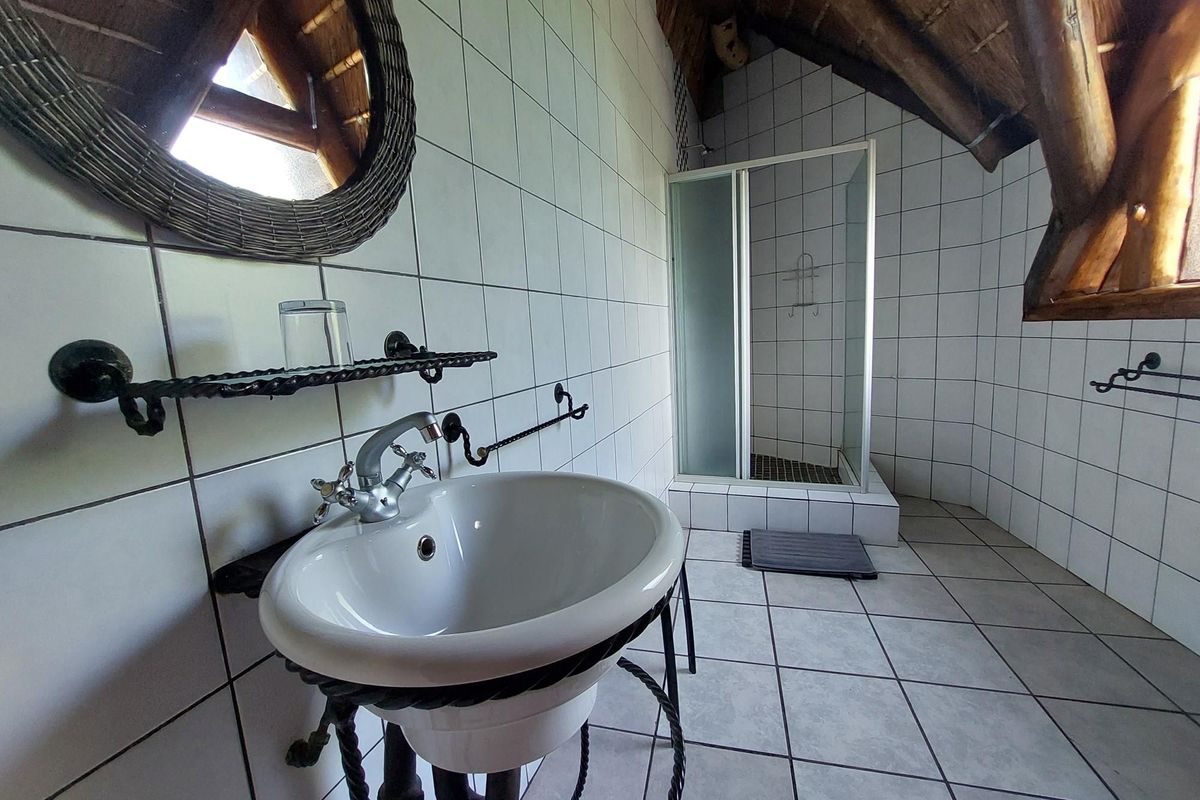 A bathroom featuring a pedestal basin, a glass-enclosed shower, and tiled walls under a rustic thatched roof structure.