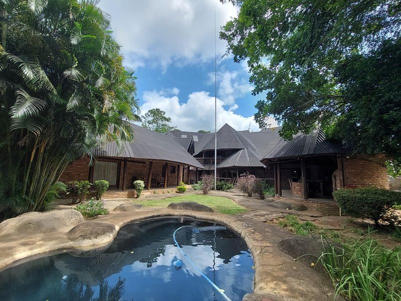 A scenic outdoor pool area featuring natural rock landscaping, a covered patio space, and a distinctive multi-pitched roof design on the main building structure.