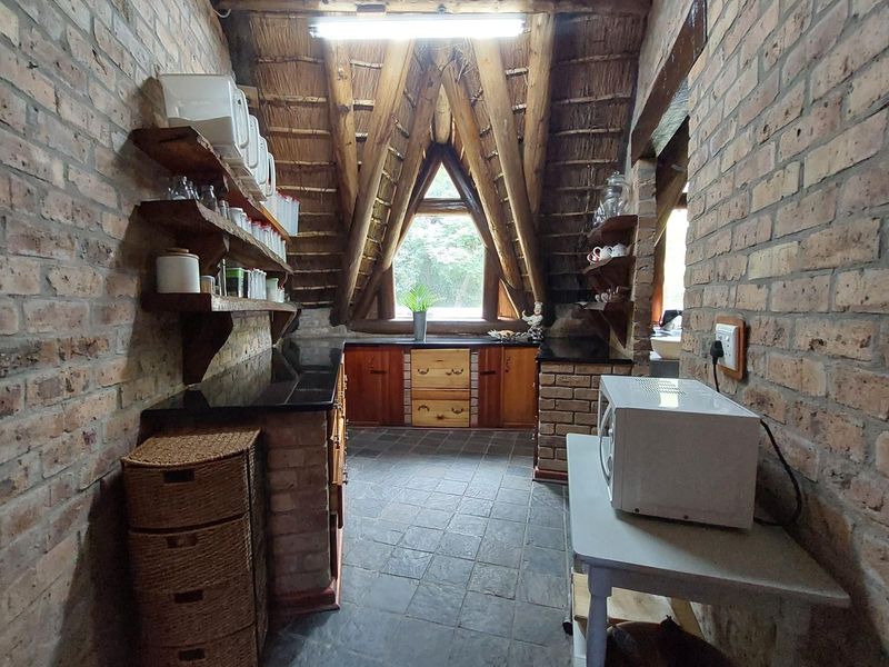 A rustic kitchen featuring exposed brick walls, a high thatched ceiling with timber beams, stone flooring, and custom built-in brick cabinetry with granite countertops.