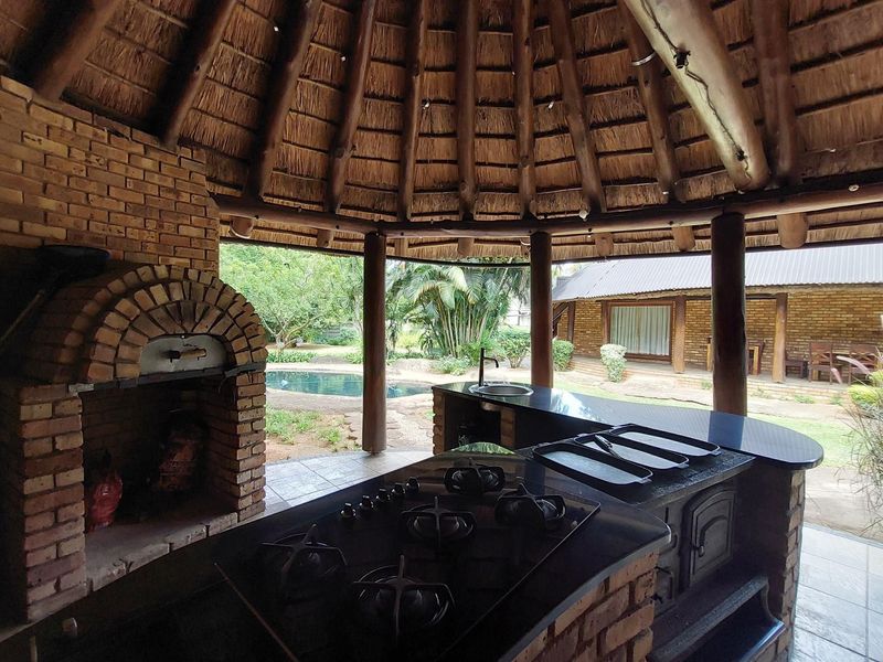 Outdoor entertainment area featuring a thatched roof, built-in brick pizza oven, gas hob, and sink with stone countertops overlooking the garden and pool.