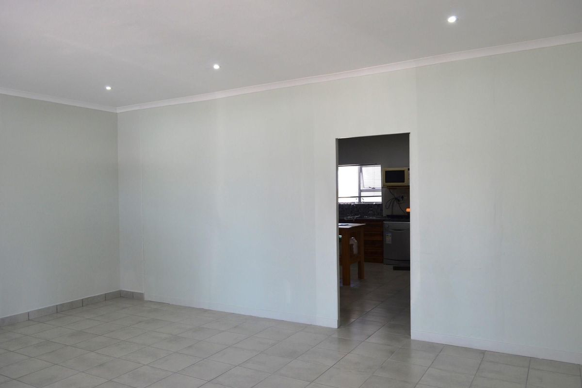 A spacious, neutral-toned living area with tiled flooring and recessed ceiling lighting, featuring a direct doorway leading into the adjacent kitchen space.