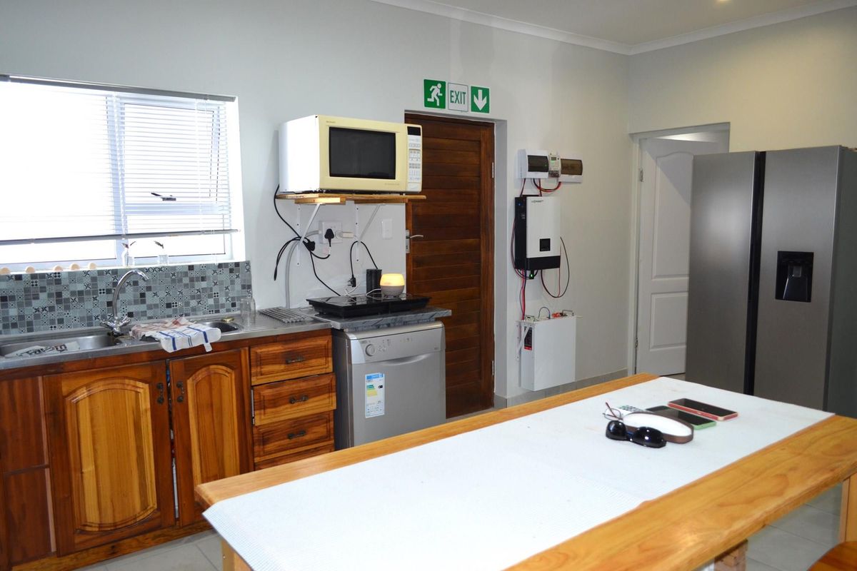A functional kitchen area featuring wooden cabinetry, a stainless steel sink, tiled backsplash, and a wall-mounted solar inverter and battery backup system.