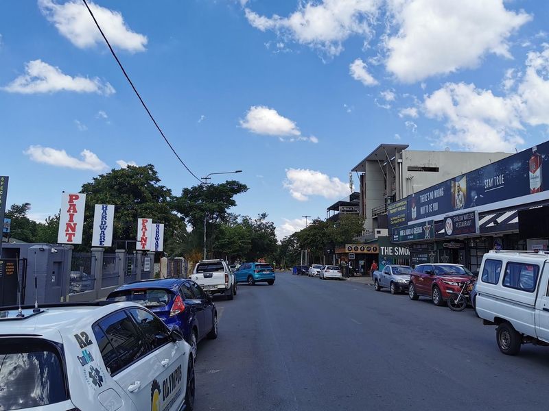 A street-level view of a commercial retail area featuring various storefronts, signage, and roadside parking along an asphalt road.