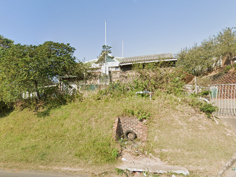 Front view of an industrial property featuring a metal security gate, concrete driveway, and significant overgrown landscaping requiring maintenance.