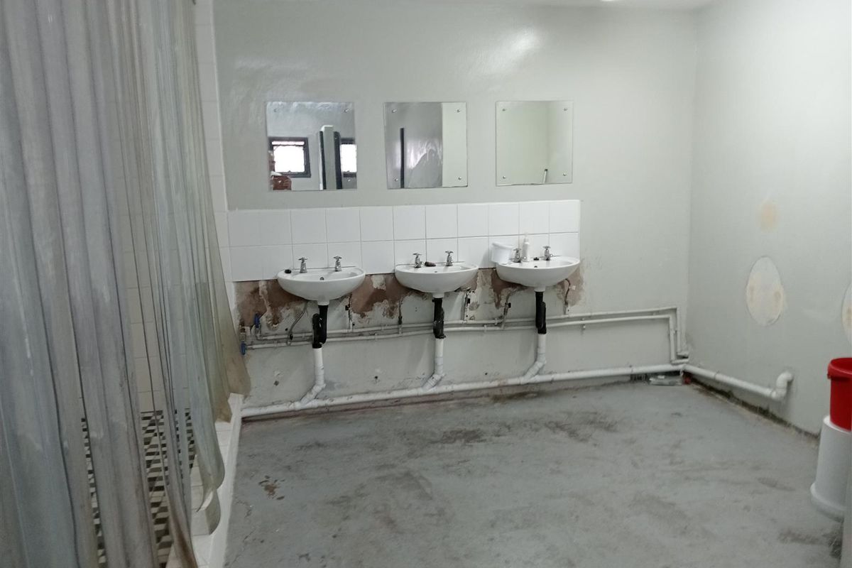 Industrial-style communal bathroom featuring three wall-mounted washbasins, exposed plumbing, and tiled splashbacks, requiring significant cosmetic renovation and wall repairs.