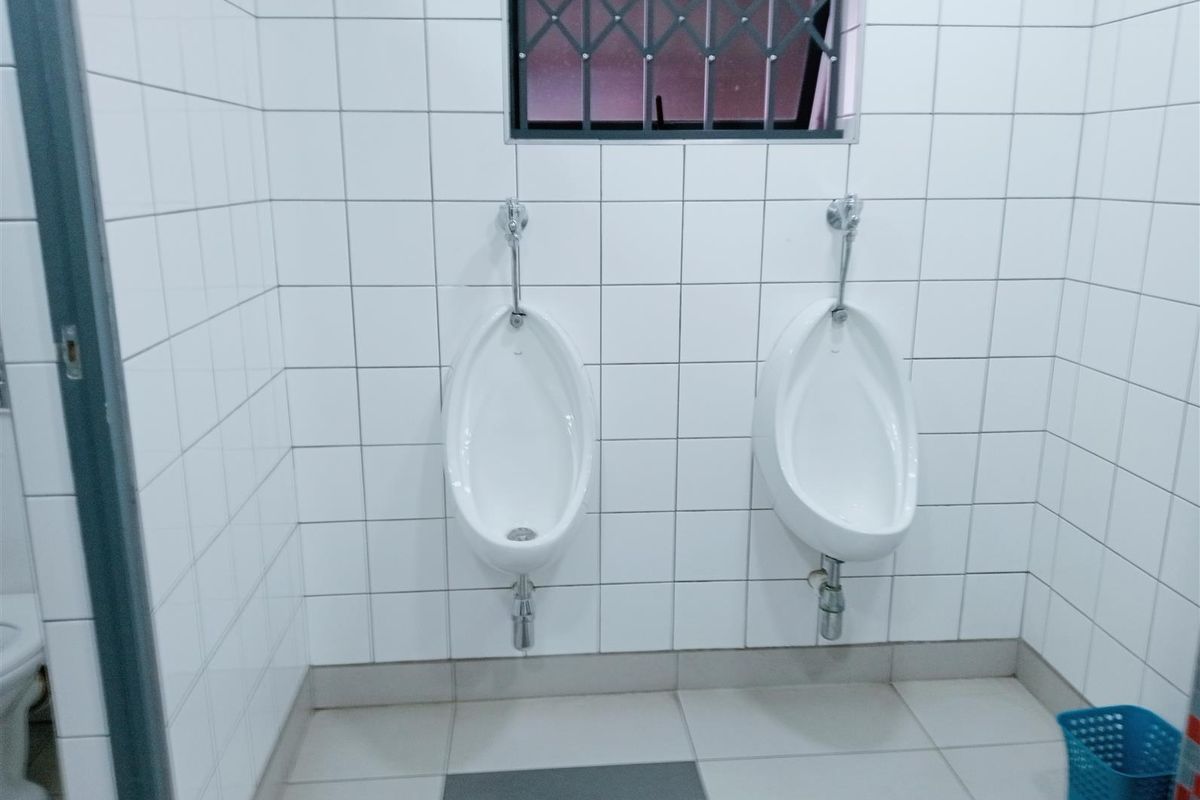 A functional industrial restroom facility featuring two wall-mounted ceramic urinals with tiled walls and flooring, including a security-grille window.