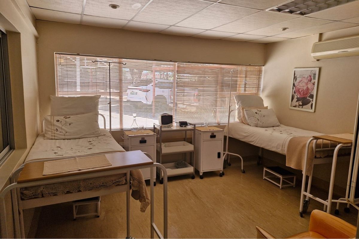 A functional shared office or medical-style room featuring vinyl flooring, suspended ceiling, air conditioning, and large windows with venetian blinds.