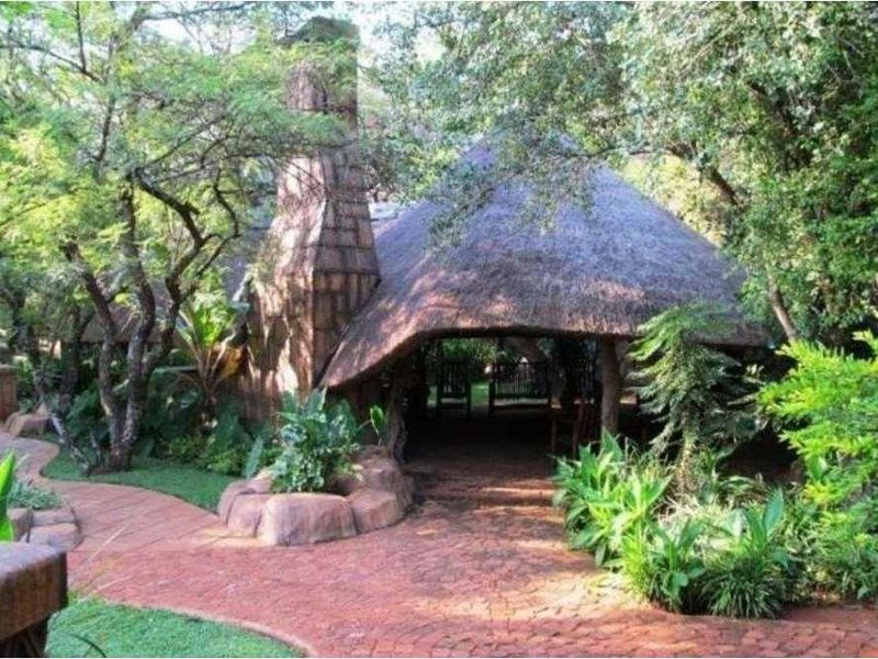 A scenic outdoor lodge area featuring a traditional thatched roof structure, a prominent stone chimney, and winding brick pathways set within lush garden surroundings.