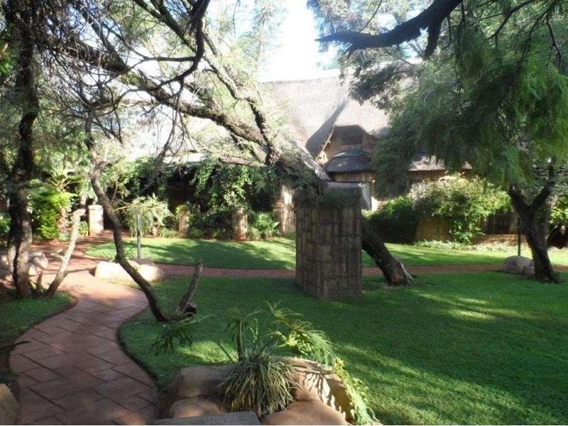 A lush, well-maintained garden featuring a paved walkway, mature trees, and stone pillars leading toward a lodge building with a traditional thatched roof.