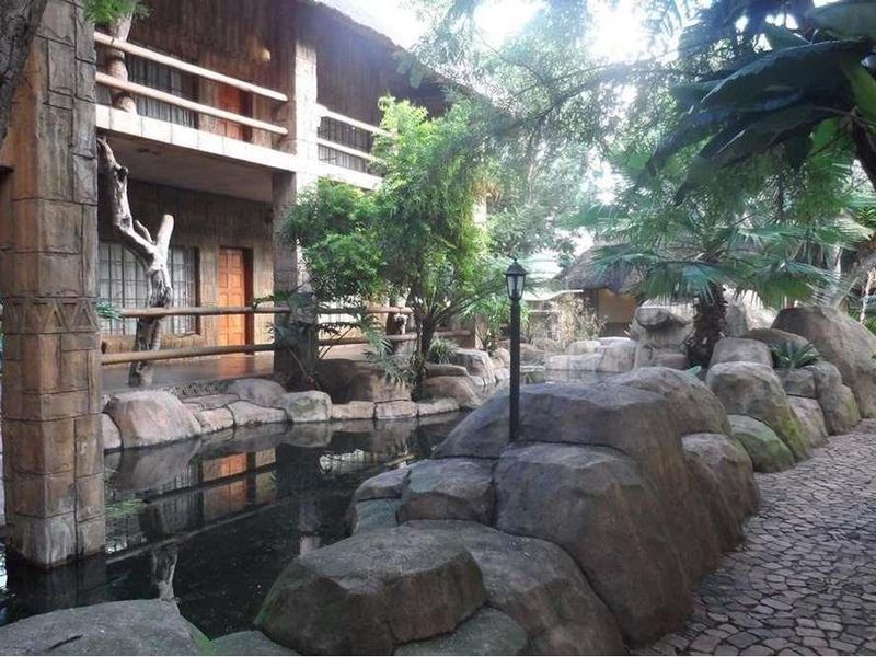 A scenic lodge exterior featuring a multi-level structure with thatched roofing, stone accents, a wooden balcony, and a landscaped water feature with stone pathways.