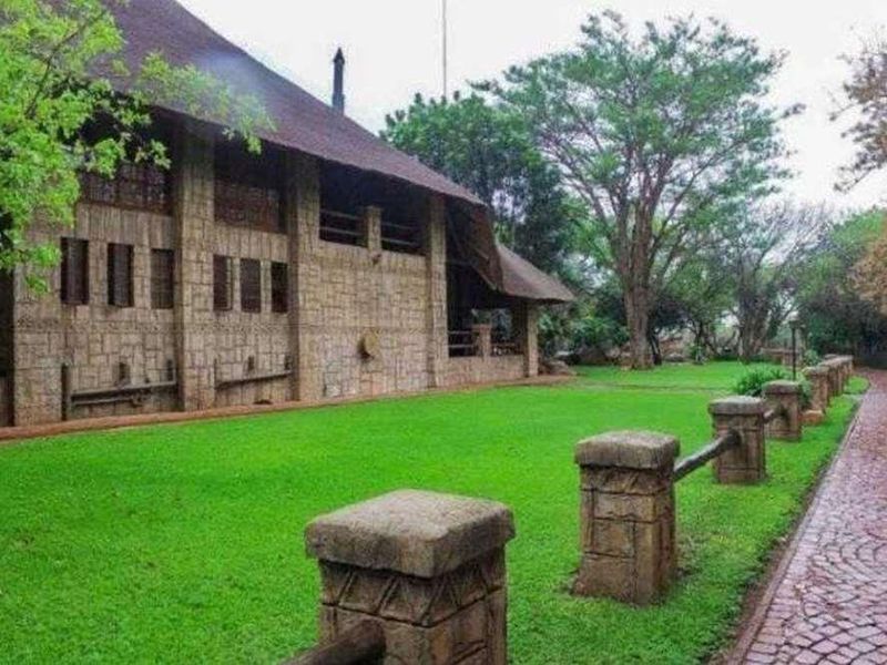 A view of the lodge exterior featuring stone cladding, a thatched roof, and a well-maintained lawn with stone pillar fencing and a cobblestone driveway.