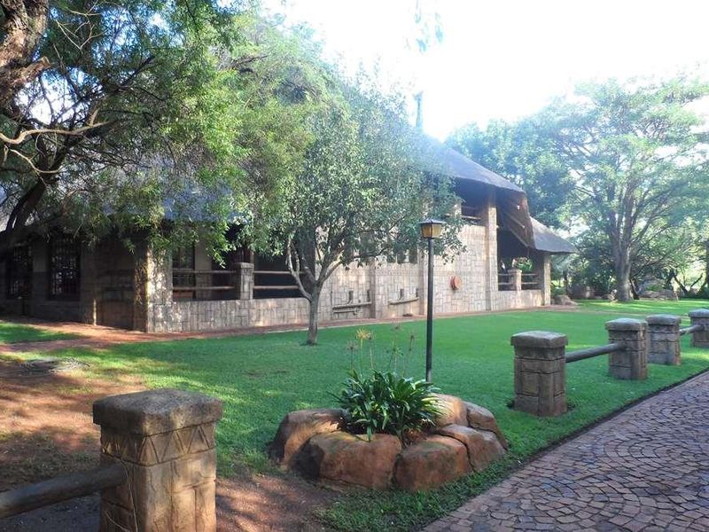 A charming lodge exterior featuring traditional stone cladding, a thatched roof, and a well-maintained garden with stone-pillared fencing and a cobblestone driveway.