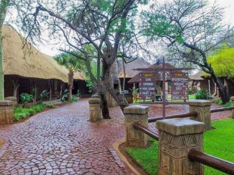 A scenic lodge exterior featuring traditional thatched roof structures, stone masonry, and a cobblestone pathway winding through a well-maintained garden setting.