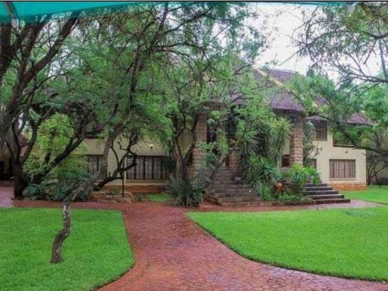 A lodge-style property featuring a stone facade, thatched roof, and a grand stone staircase leading to the entrance, surrounded by a lush garden.