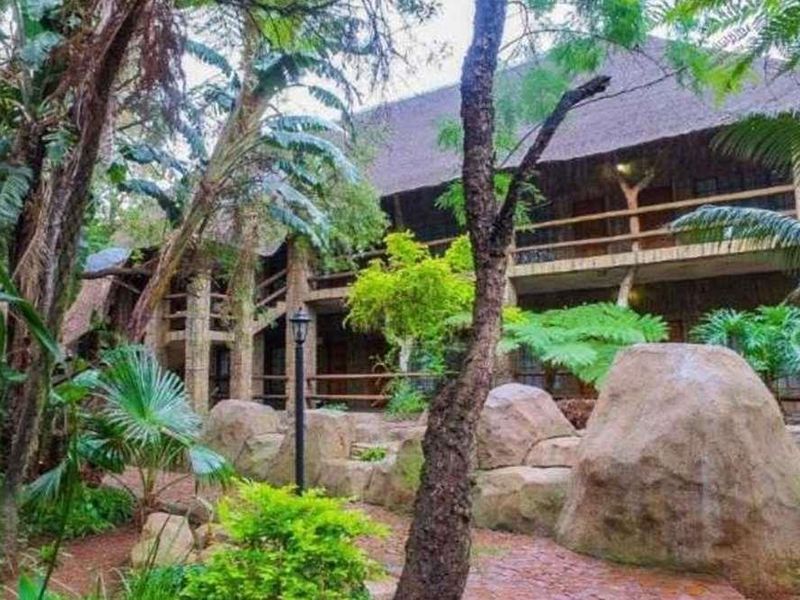 A multi-story lodge exterior featuring traditional thatch roofing, timber balcony railings, and natural stone landscaping in a lush garden setting.