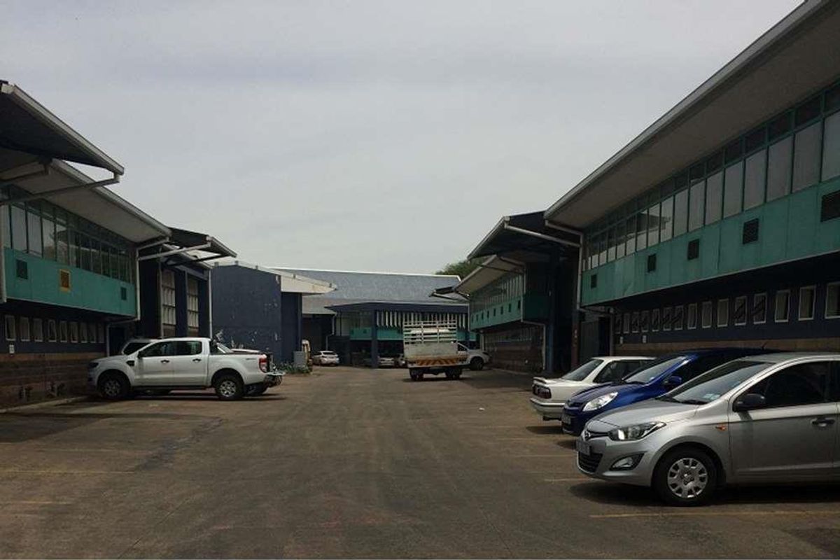 Exterior view of an industrial complex featuring multiple warehouse units with metal roofing, clerestory windows, and a central paved parking area.
