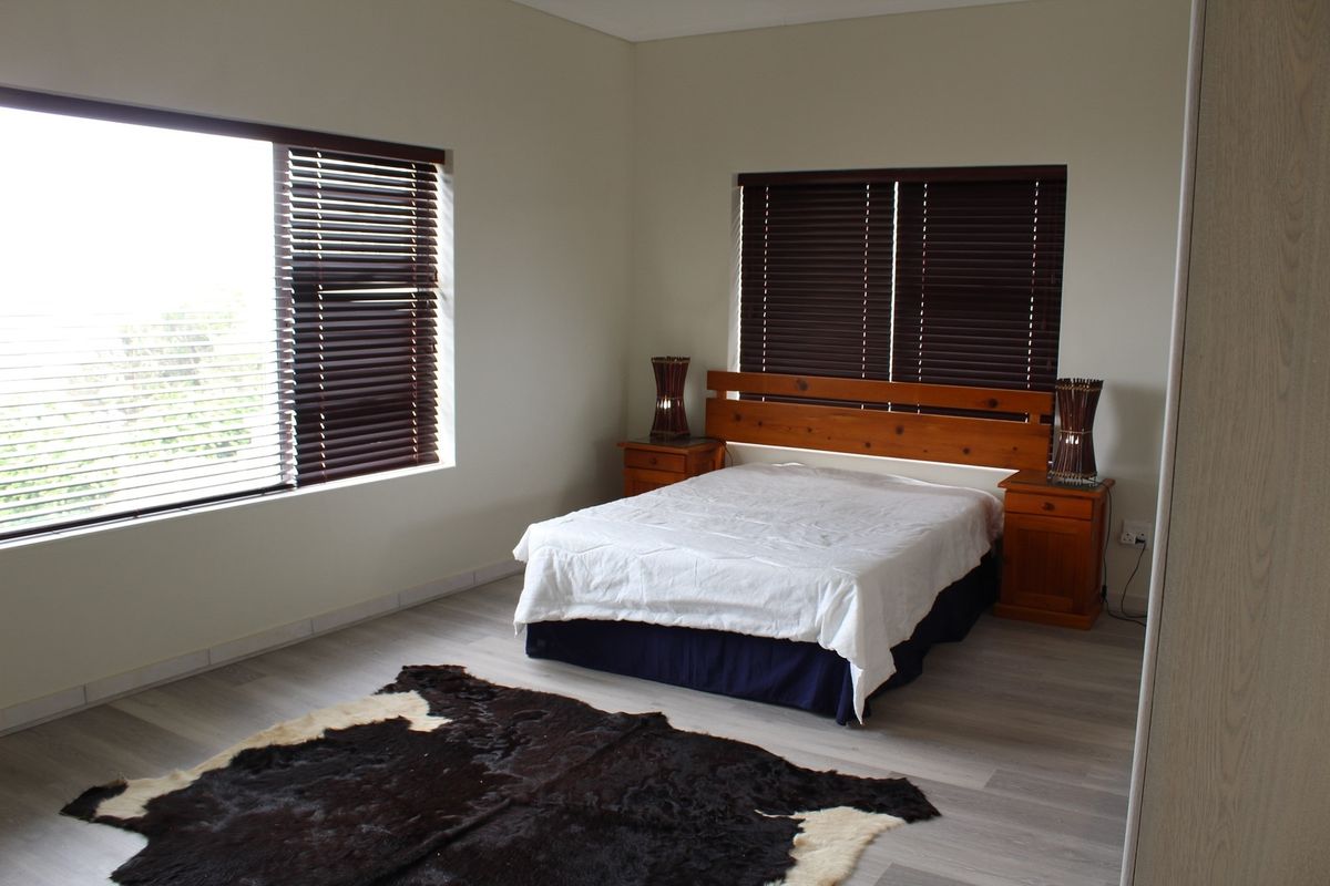 A spacious bedroom featuring light-toned laminate flooring, neutral walls, and large windows fitted with dark timber venetian blinds.