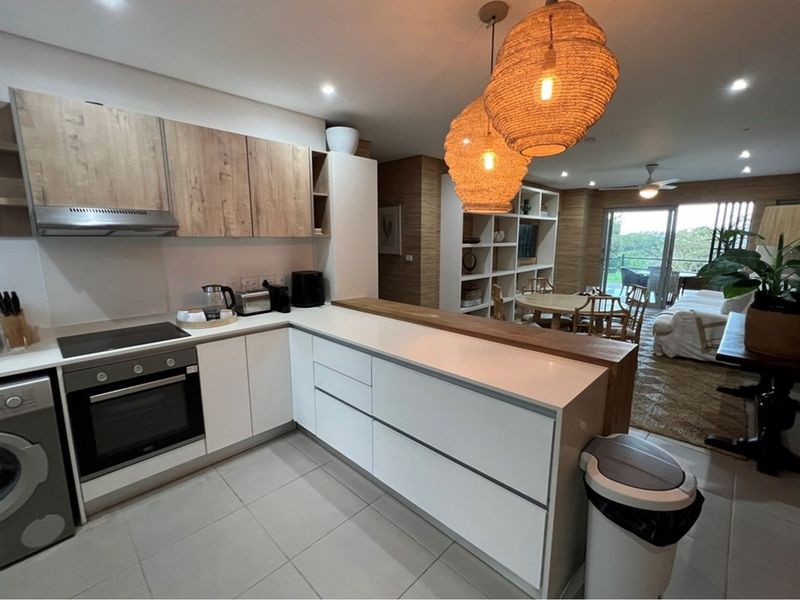 Modern open-plan kitchen featuring sleek white and wood-grain cabinetry, stone countertops, and a breakfast bar, seamlessly flowing into the adjacent living and dining area.
