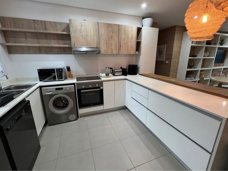 Modern kitchen featuring sleek white cabinetry, wood-tone accents, stone countertops, integrated oven and hob, and a convenient breakfast bar overlooking the living area.