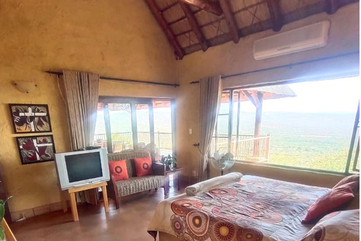 A rustic bedroom featuring a high thatched ceiling, textured walls, and large windows offering expansive views, complemented by an integrated air conditioning unit.