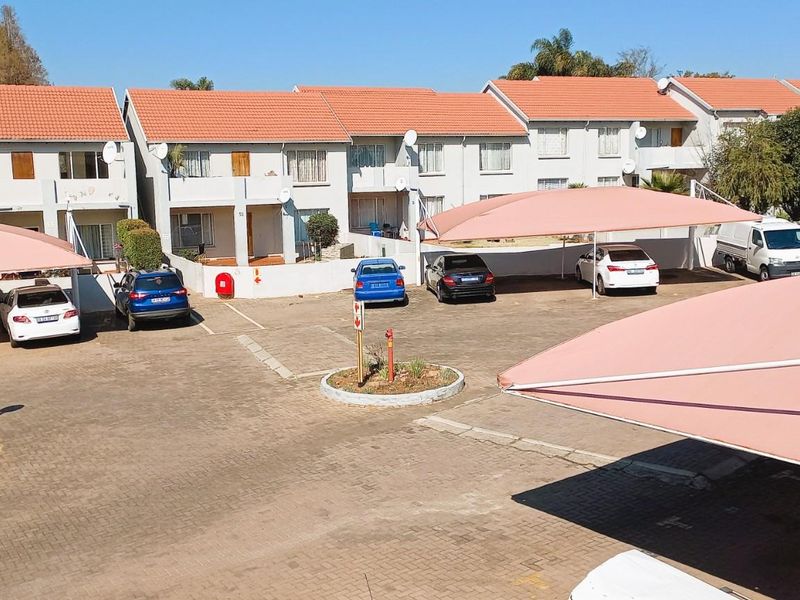 Exterior view of a townhouse complex featuring a paved parking area with shade cloth carports and multi-story residential units with tiled roofs.
