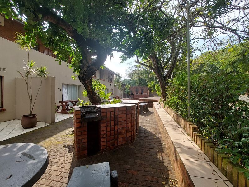 Outdoor communal entertainment area featuring a built-in brick braai, paved patio space, and established trees within a residential complex setting.