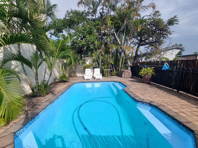 A private outdoor swimming pool area featuring a paved deck, boundary fencing, and established tropical landscaping.