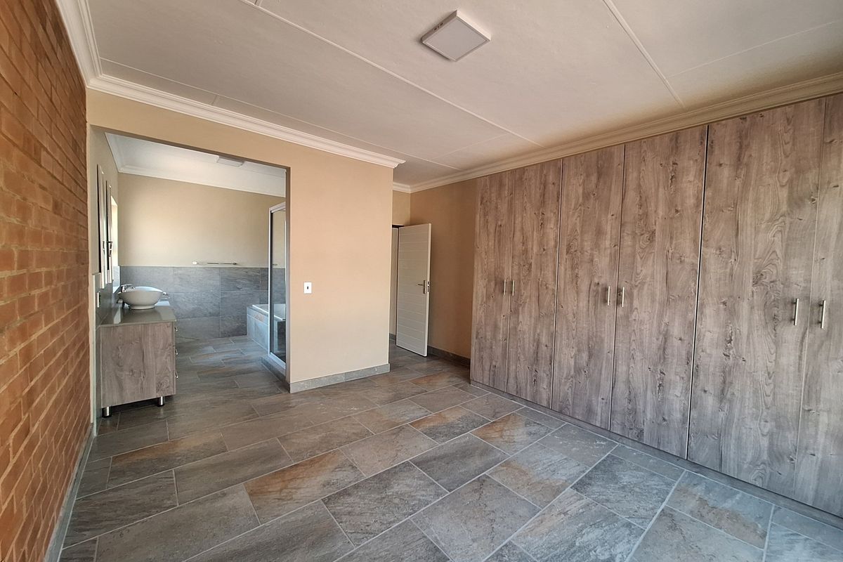 Spacious bedroom featuring built-in wood-look cabinetry, tiled flooring, and an exposed brick feature wall, with an adjoining en-suite bathroom.