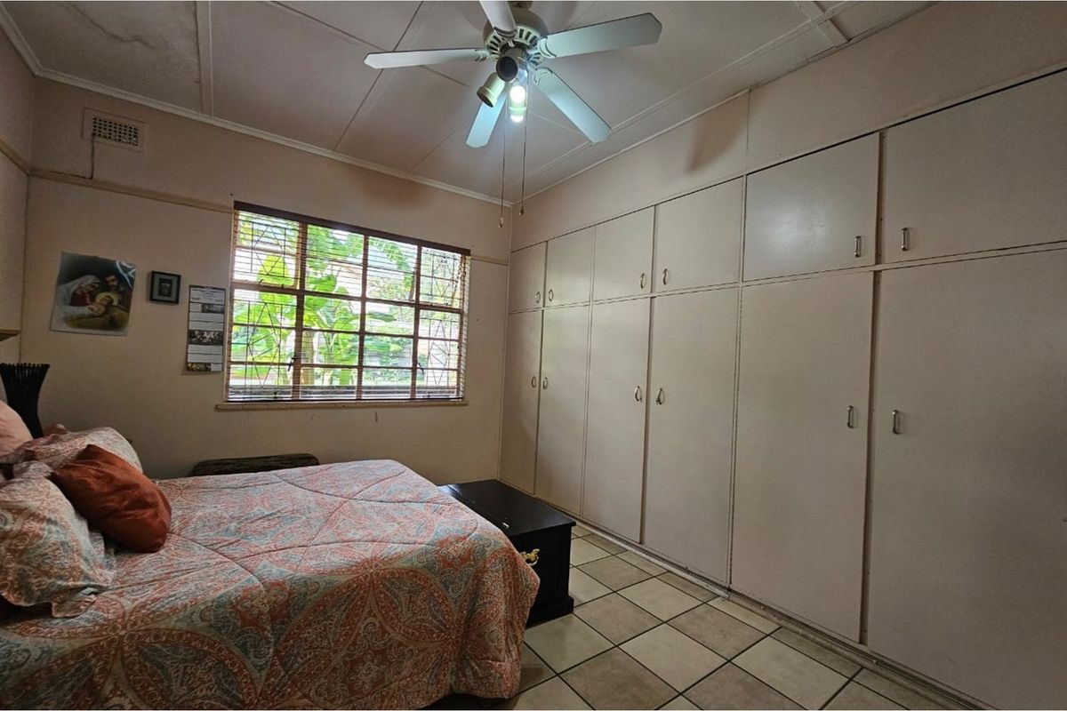 A bedroom featuring built-in floor-to-ceiling wardrobes, tiled flooring, a ceiling fan, and a window with security bars.