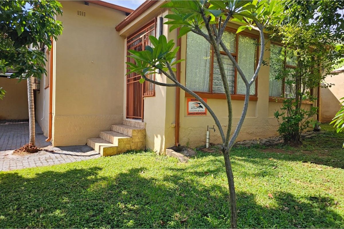 Exterior view of the side entrance featuring a paved walkway, small concrete steps, and established garden area with security signage.