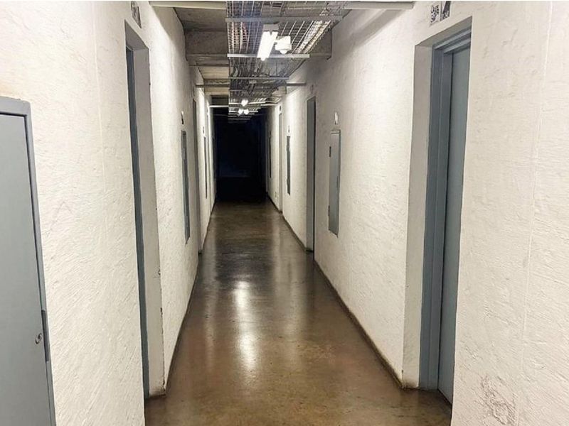 A utilitarian apartment building hallway featuring exposed industrial conduit, concrete flooring, and multiple access doors.