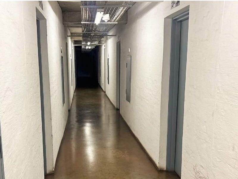 A utilitarian apartment hallway featuring exposed conduit, concrete flooring, and multiple access doors, presenting a basic industrial aesthetic.