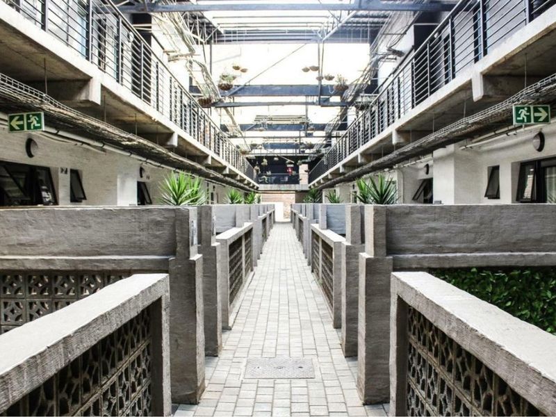 An industrial-style open-air apartment complex corridor featuring concrete walkways, breeze block partitions, and an exposed steel roof structure.