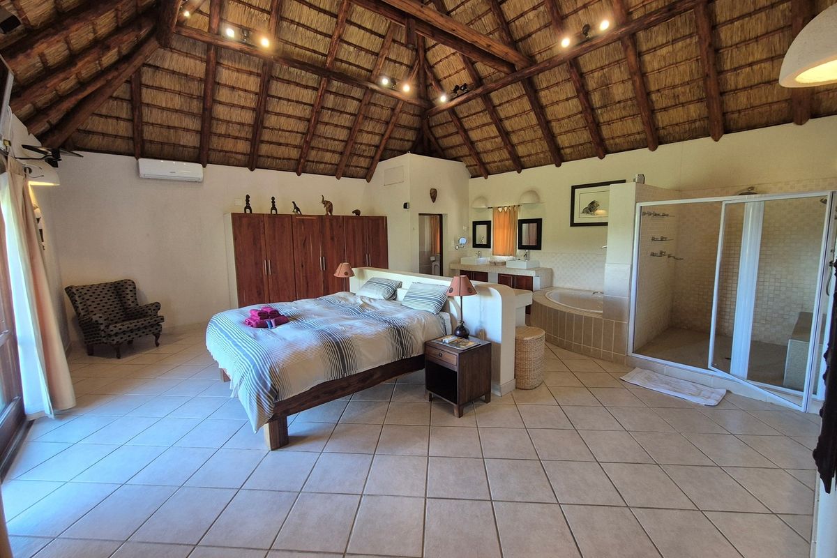 Spacious open-plan bedroom featuring a high thatched roof, tiled flooring, built-in cabinetry, and an integrated en-suite bathroom with a shower and soaking tub.