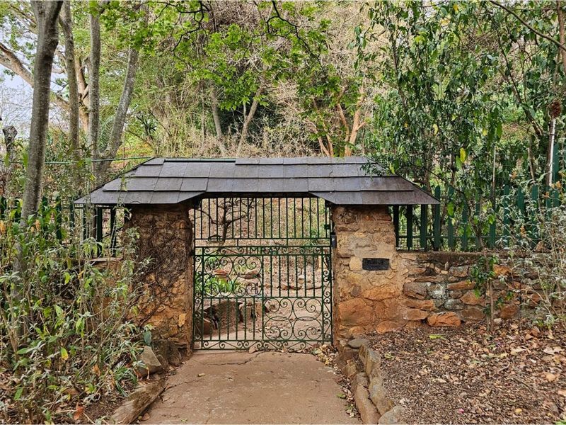 A charming stone-walled garden entrance featuring a decorative wrought iron gate and a small pitched roof structure, set within a lush, mature garden.