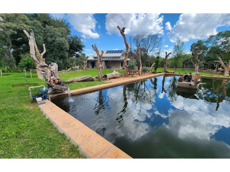 A tranquil garden setting featuring a large swimming pool with a water feature, surrounded by landscaped lawns and mature trees.