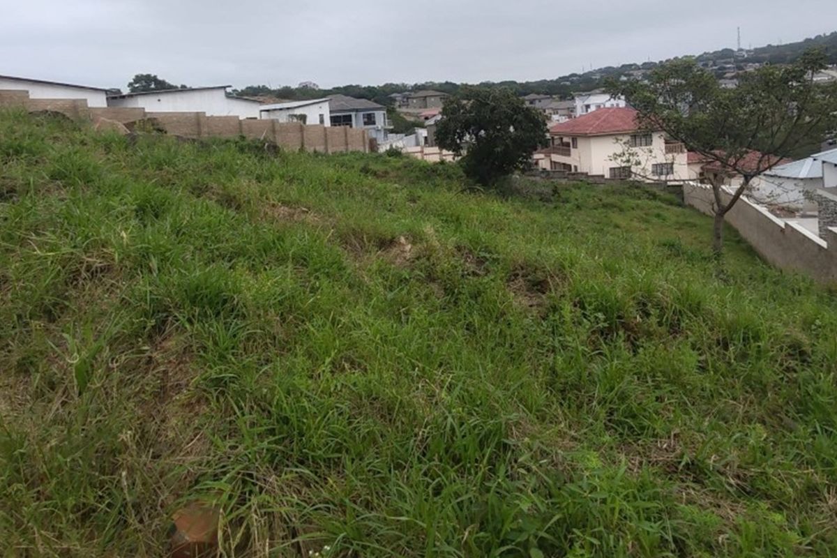 A sloped, undeveloped residential plot covered in natural grass, featuring perimeter boundary walls and views of the surrounding neighborhood.
