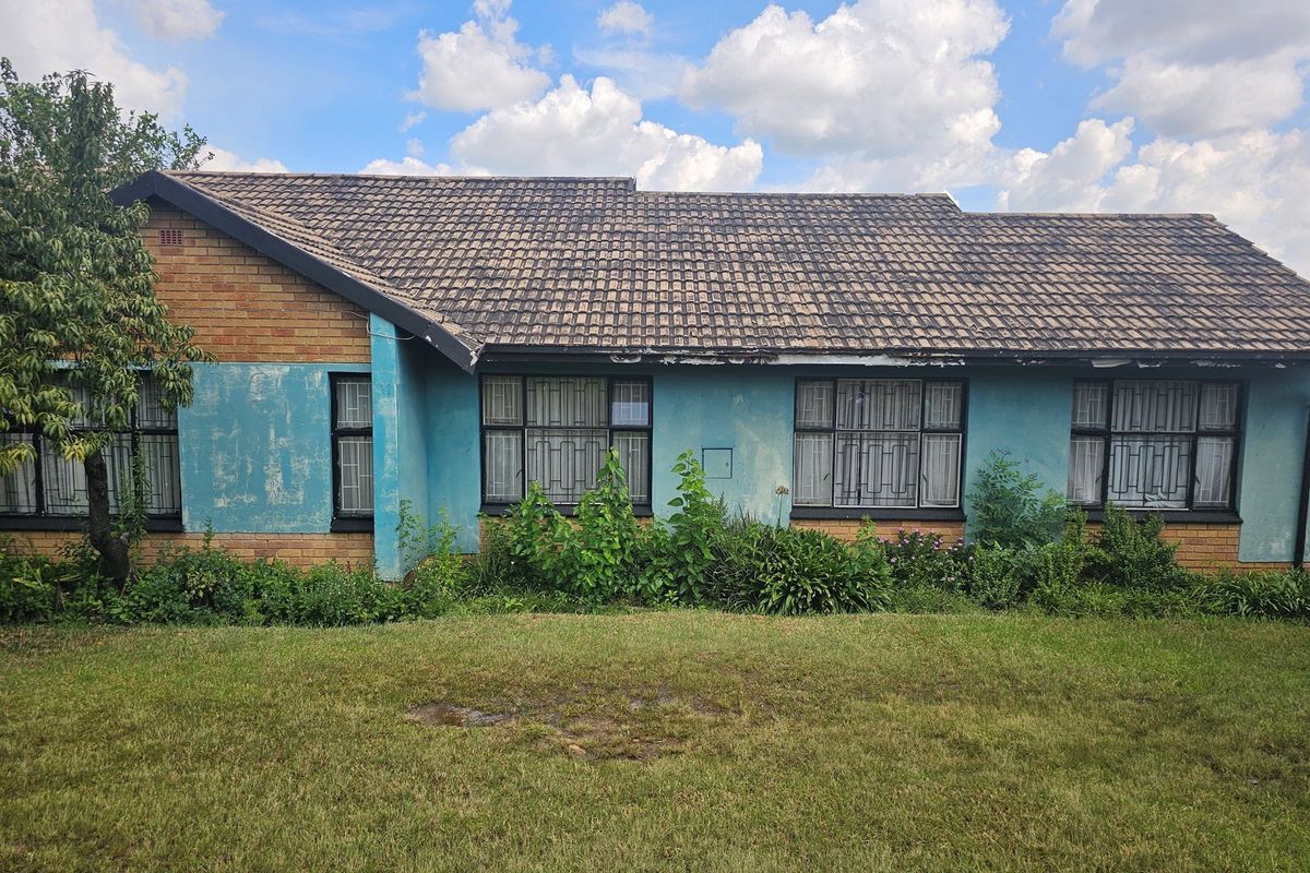 Rear view of a single-story brick house featuring a tiled roof, security-barred windows, and a garden area requiring maintenance and exterior paint restoration.