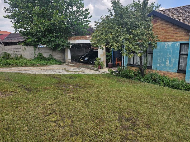 Front exterior view of a brick house featuring a carport, large lawn area, and boundary wall, showing signs of wear and maintenance requirements.